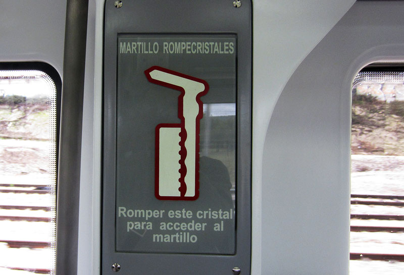 Foto de un paradójico sistema de seguridad en trenes de Cercanías de Renfe, que reza: «Martillo rompecristales: romper este cristal para acceder al martillo».